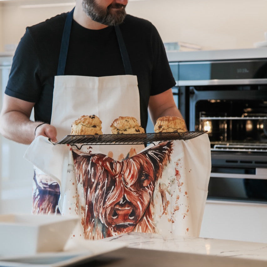 Man in a kitchen wearing an apron with a cow design, holding a tray of scones.